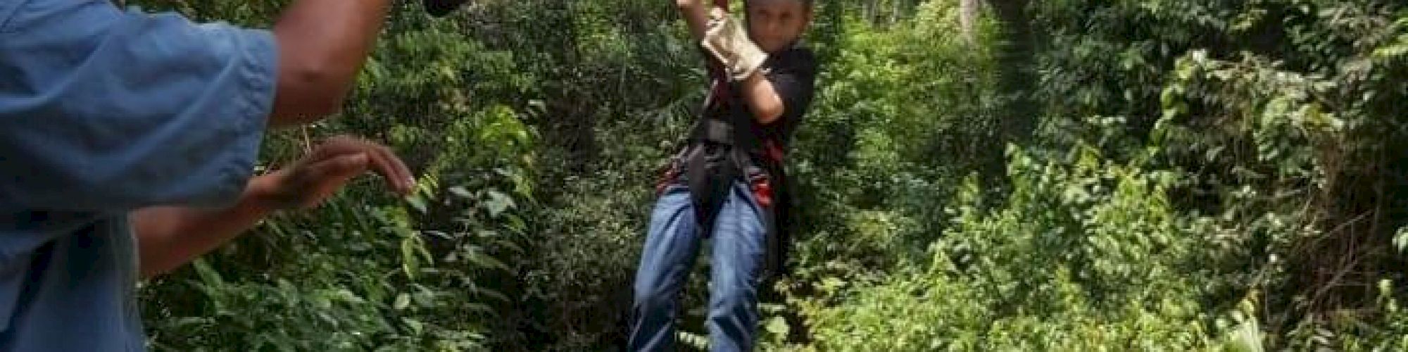 An individual is ziplining through a forested area while another person on the ground appears to be assisting or observing.