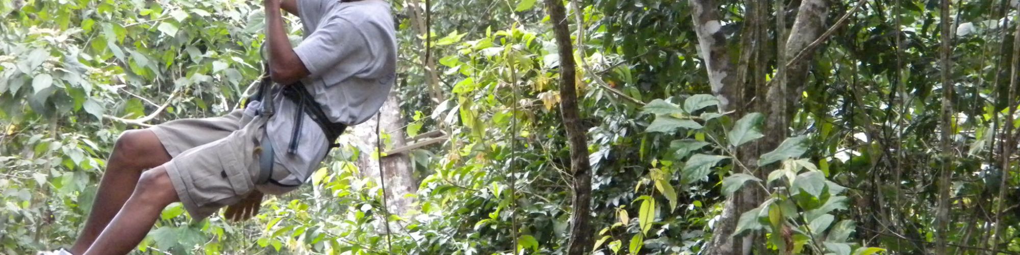 A person wearing a red helmet rides a zipline through a lush green forest, smiling and enjoying the adventure.