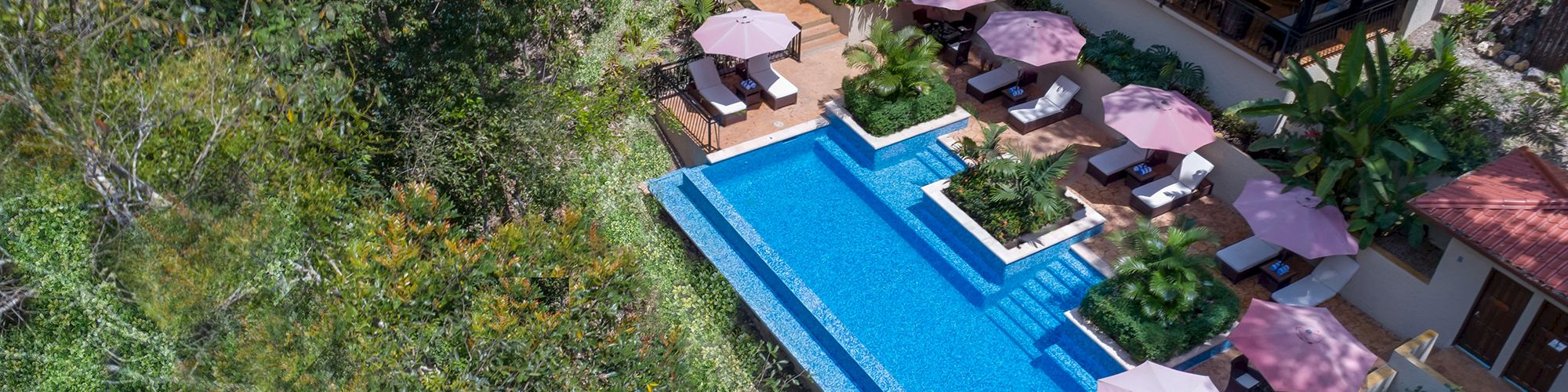 The image shows an aerial view of a pool area with lounge chairs, umbrellas, greenery, and a building with a red roof in a forested setting.