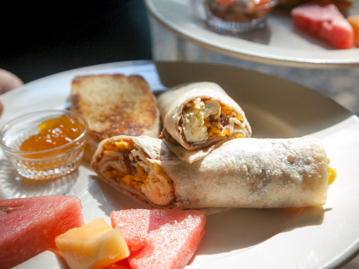 The image shows a plate with a breakfast burrito cut in half, toast, a small dish of jam, and watermelon chunks, with another plate holding more fruit.