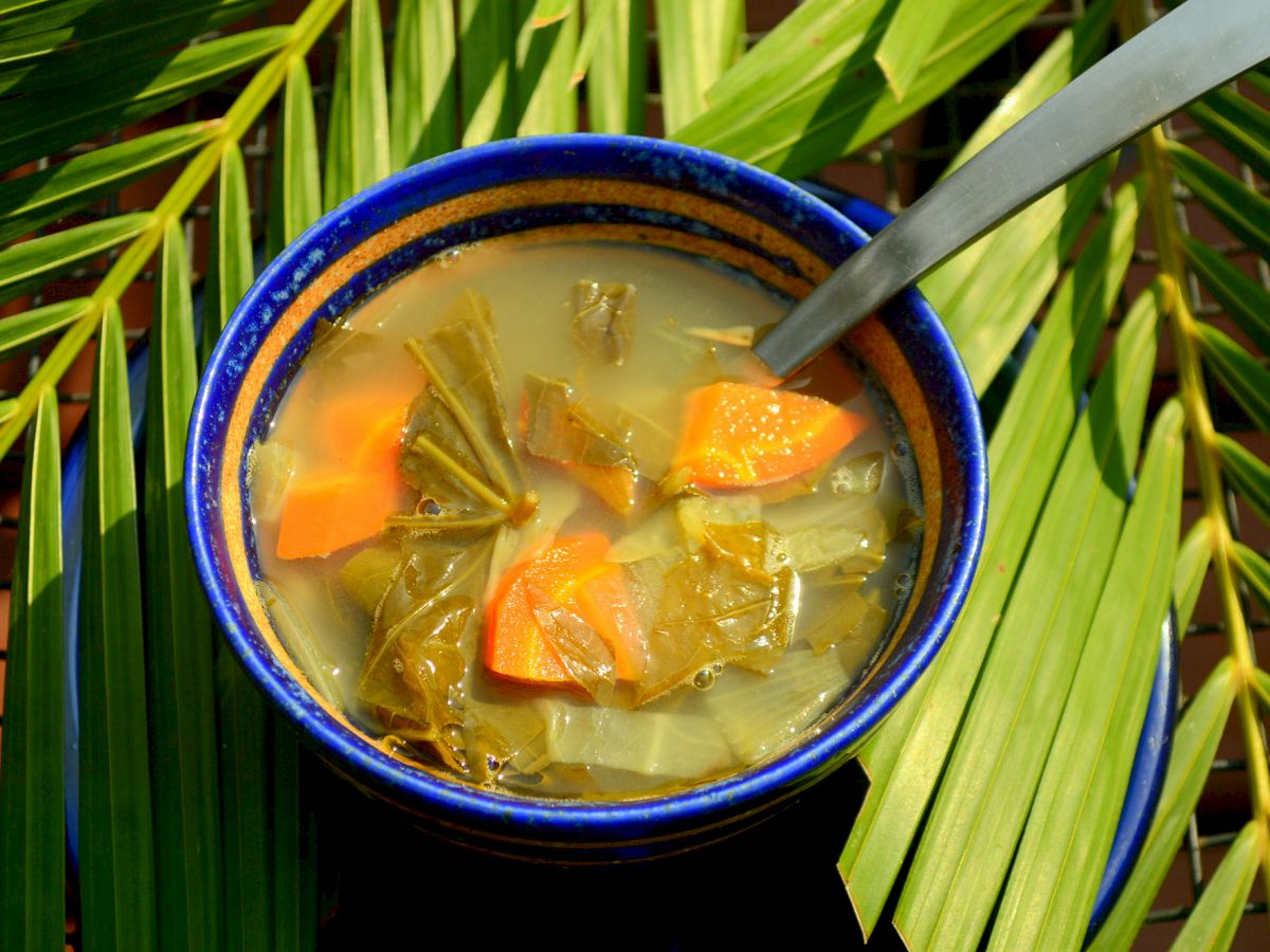 A bowl of vegetable soup with leafy greens and chunks of orange vegetables is placed on green palm leaves with a spoon in the soup.