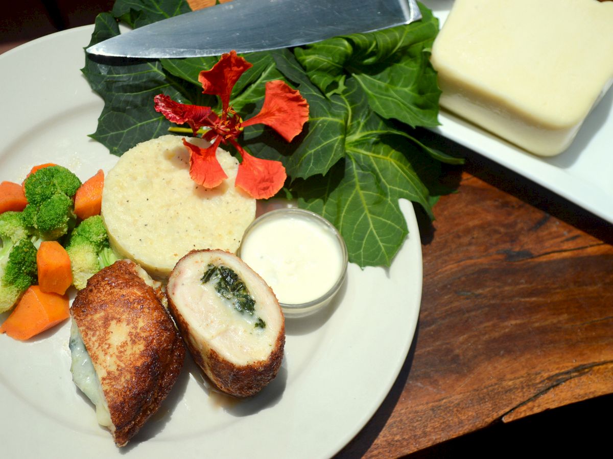 The image shows a plate with stuffed chicken, a sauce, a flatbread, and mixed vegetables, along with a side display of leafy greens and cheese blocks.