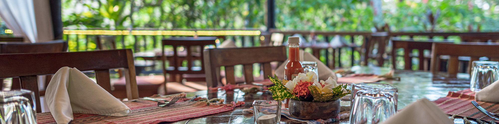 This image shows a dining table set in an outdoor restaurant with napkins, glasses, and table decorations, surrounded by lush greenery.