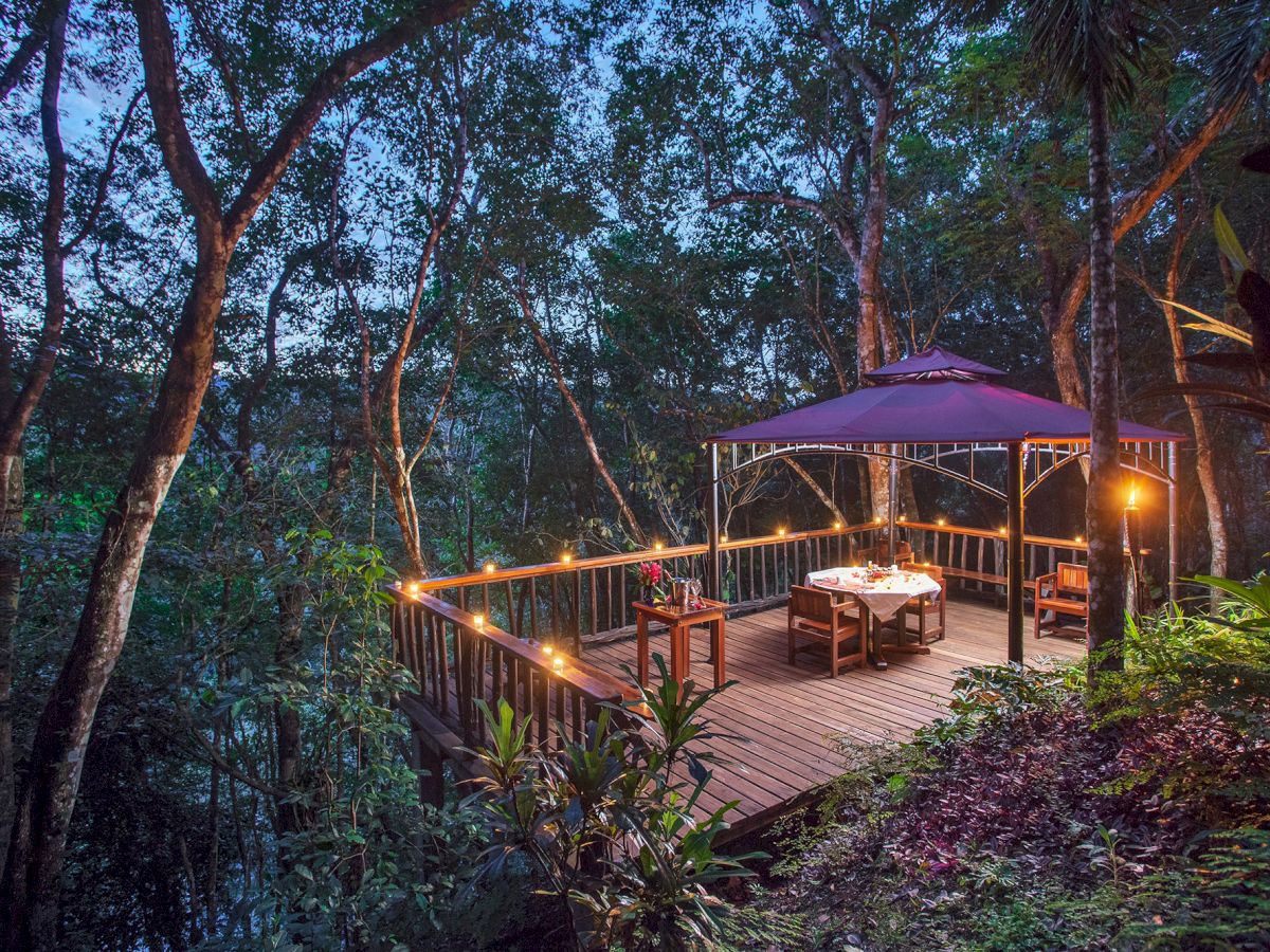 A romantic, lantern-lit dining setup on a wooden deck in a forested area, featuring a table with chairs under a purple canopy in the evening.