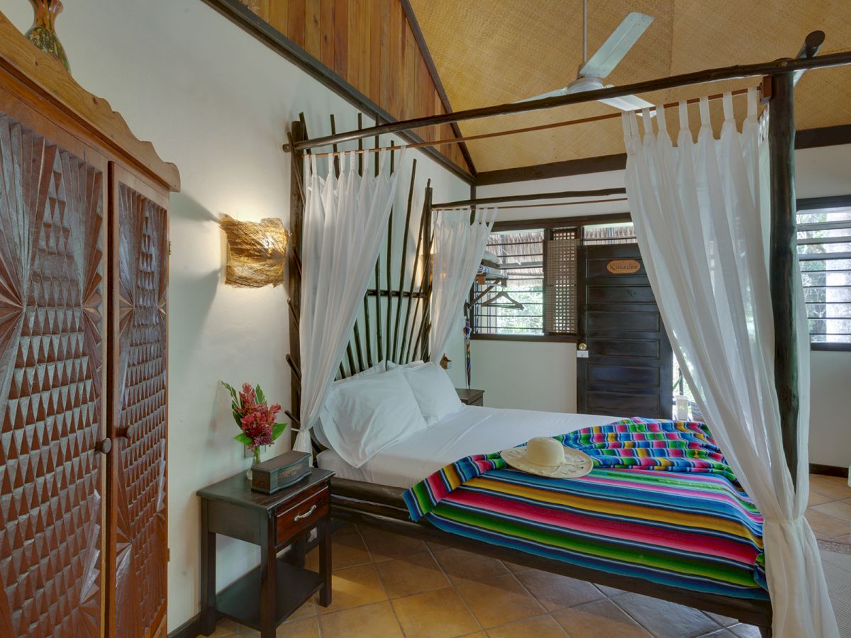 A cozy bedroom with a canopy bed adorned with white curtains, colorful striped blanket, wooden furniture, and fresh flowers in a vase.