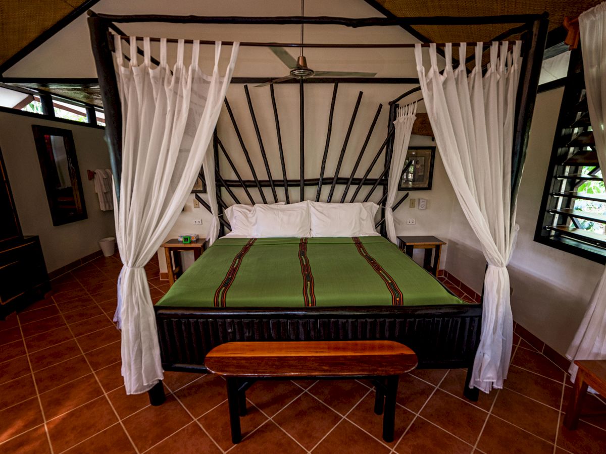 A cozy bedroom with a canopy bed, green bedspread, white curtains, wooden bench, and tiled floor, featuring rustic decor and natural light.