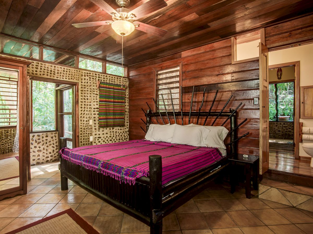 Rustic bedroom with a wooden bed, vibrant purple blanket, tiled floor, wood-paneled walls, ceiling fan, and adjoining bathroom.