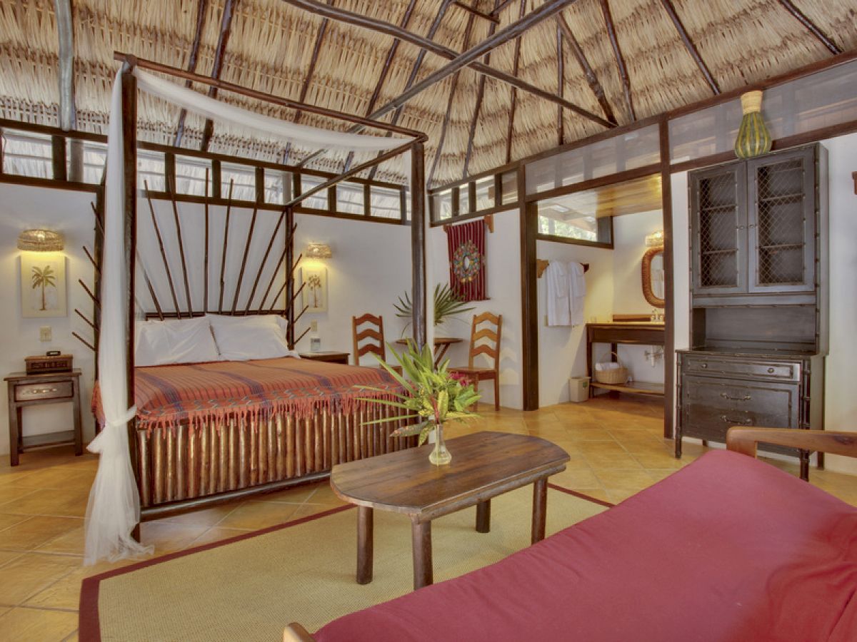 A tropical-style bedroom with a thatched roof, a canopy bed, wooden furniture, a small coffee table, and a bench, decorated with plants and bamboo.