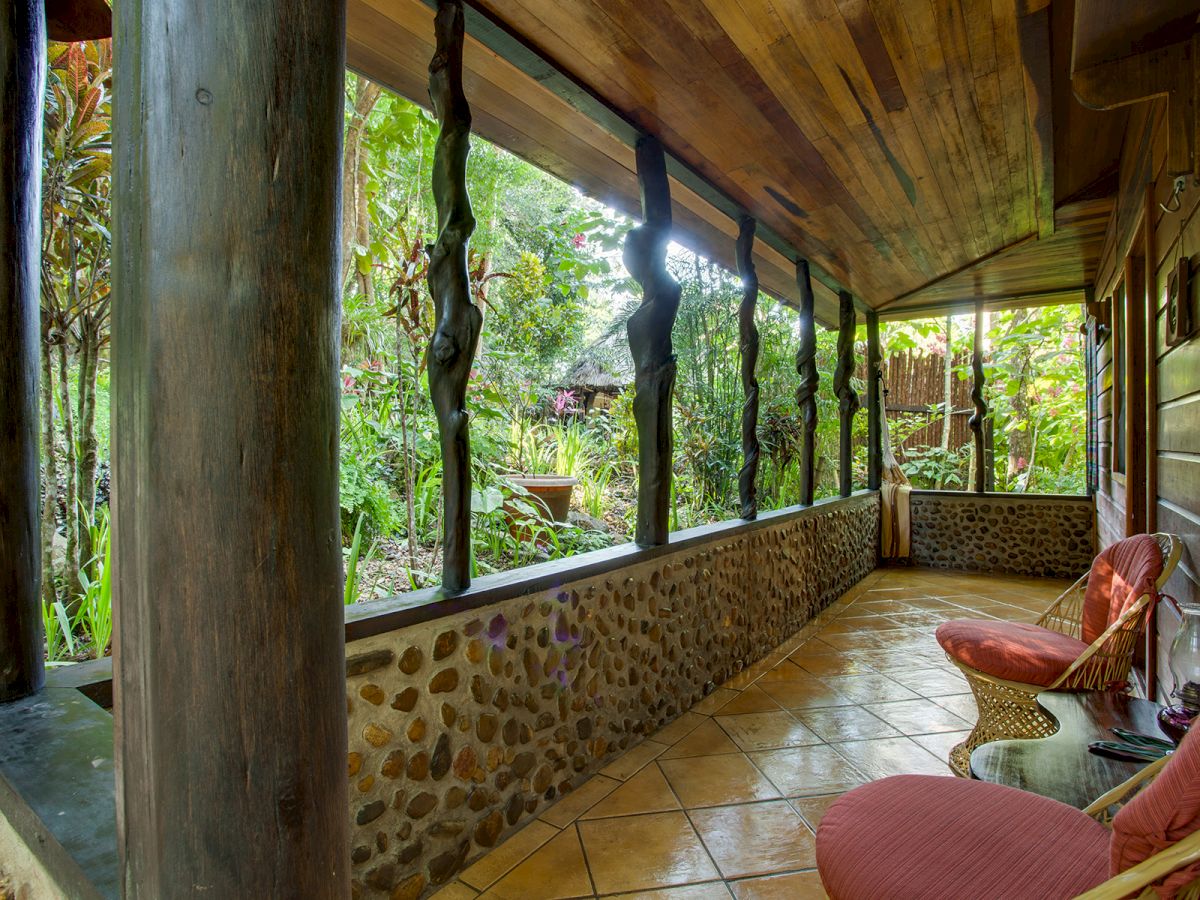 A cozy, rustic veranda with wicker chairs, wooden columns, and a stone wall overlooks a lush, green garden. The ceiling and floor are wooden.