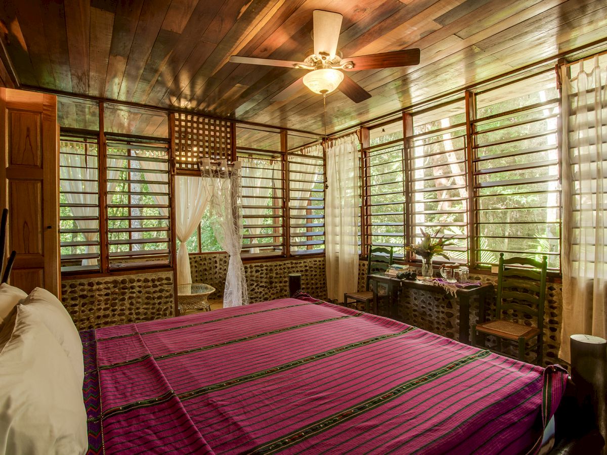 A cozy bedroom with a wooden ceiling, a large bed with a purple cover, large windows with blinds, and a ceiling fan.