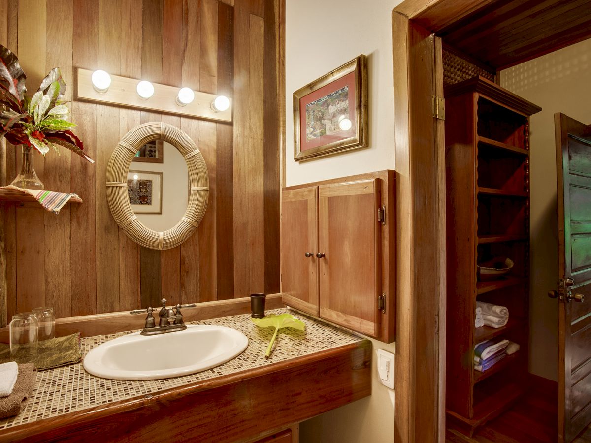 A rustic bathroom features a sink, a circular mirror, and wooden cabinets, with an open doorway leading to a towel shelf and partially visible greenery.