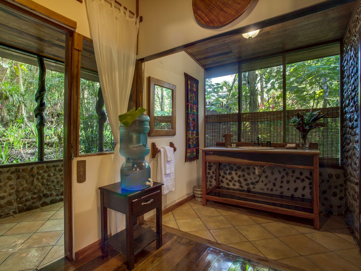 A bathroom with large windows showcasing lush greenery, a sink, a water cooler, a mirror, and tiled floors. The space is well-lit and serene.