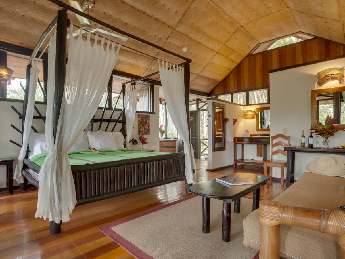 A cozy bedroom with a four-poster bed, wooden furniture, a sofa, a desk, and a coffee table, featuring large windows and natural light ending the sentence.