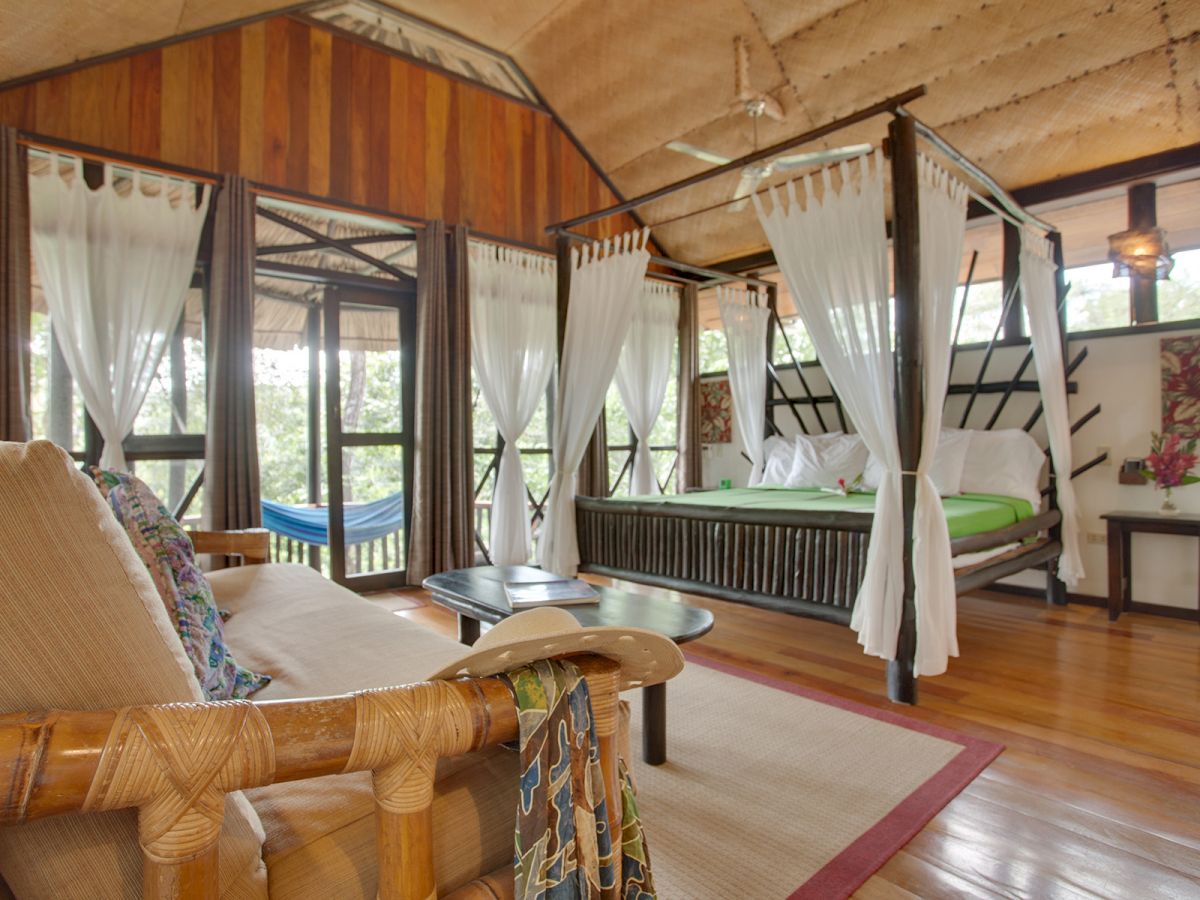 The image shows a cozy room with a four-poster bed, a wooden sofa, large windows, and a hammock outside, creating a serene and inviting atmosphere.