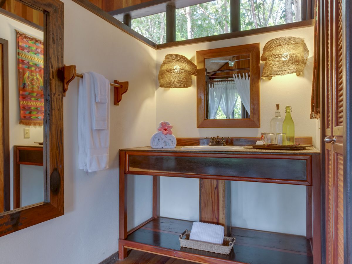 A cozy bathroom with wooden accents, featuring a mirror, sink, wall lights, and towels. Warm tones and natural light give a rustic vibe.