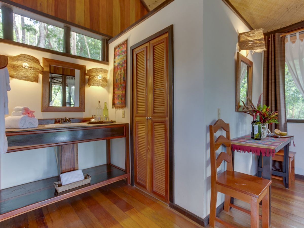 This image shows a cozy room with wooden furnishings, including a sink area with a mirror, a desk with a chair, and a window with curtains.