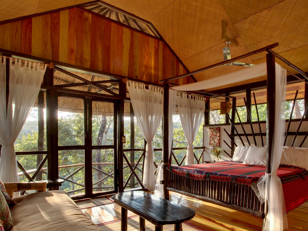 A cozy, rustic bedroom with large windows, a four-poster bed draped with white curtains, and wooden furniture, all overlooking a lush forest.