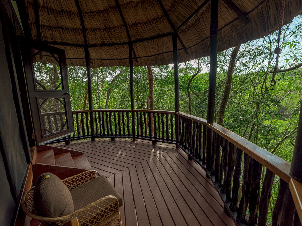 A cozy balcony with wooden flooring and railings, surrounded by lush greenery, featuring an armchair for relaxation, under a thatched roof.