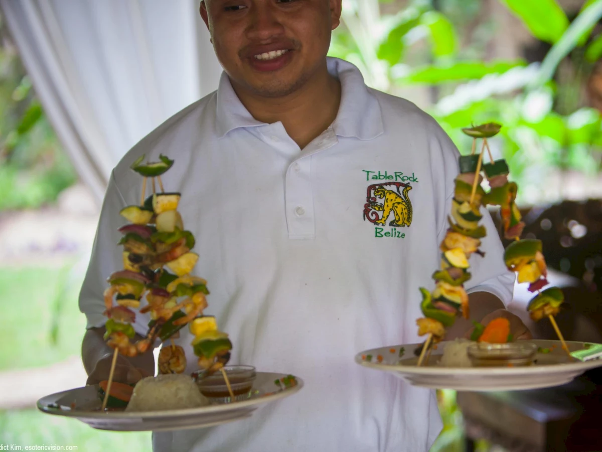 A person in a white shirt, holding two plates with skewered food at Table Rock Belize, photographed in an outdoor setting with greenery in the background.