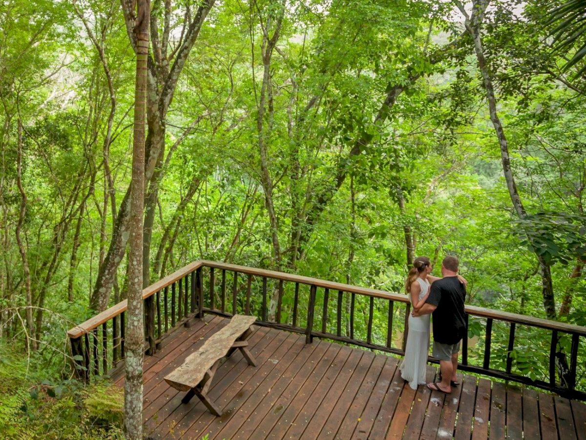 A couple stands on a wooden deck in the middle of a lush, green forest, surrounded by tall trees and dense foliage, enjoying a serene moment.