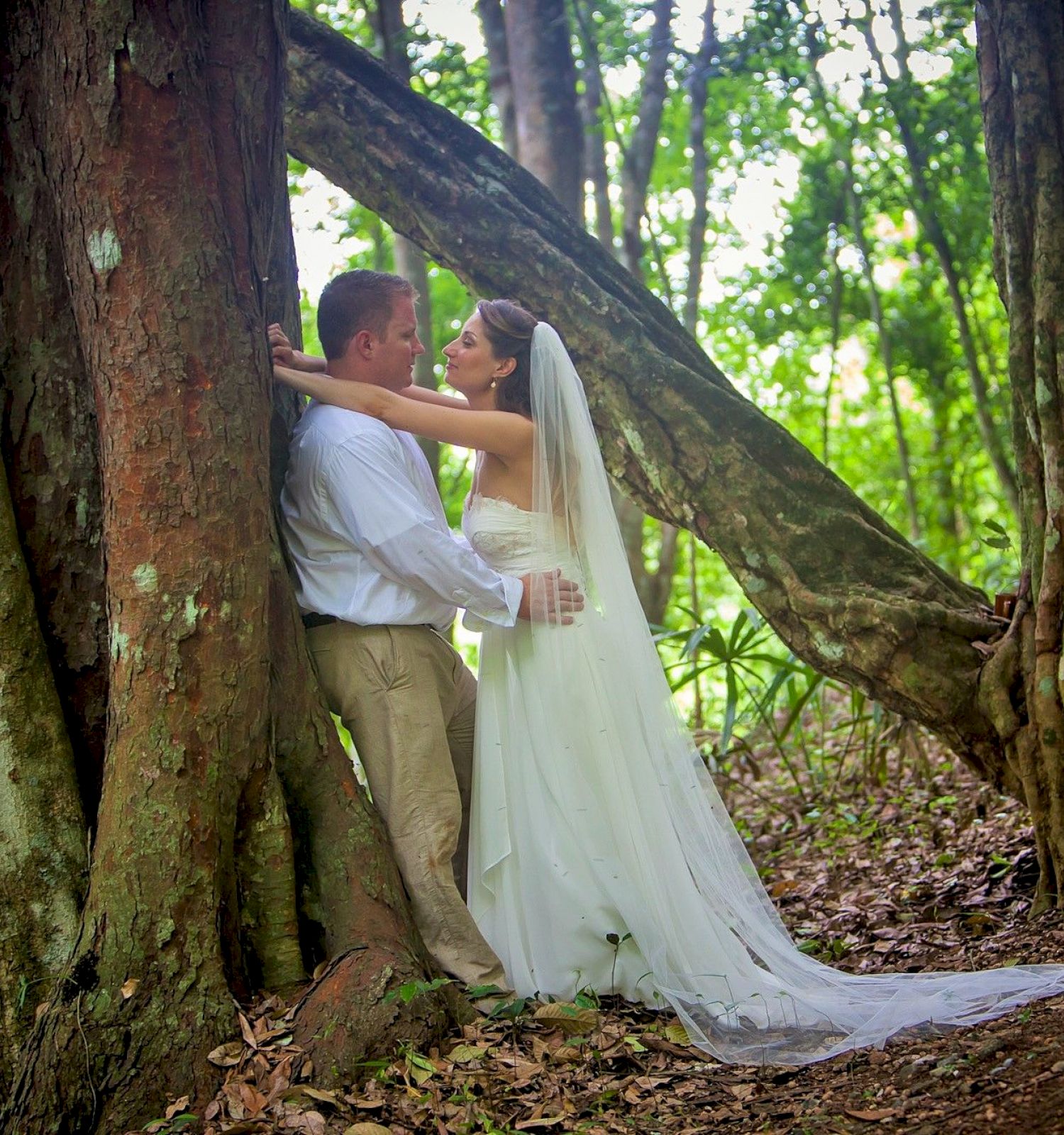 A bride and groom are embracing next to a tree in a forest, with the bride wearing a white dress and veil, and the groom in a white shirt and beige pants.