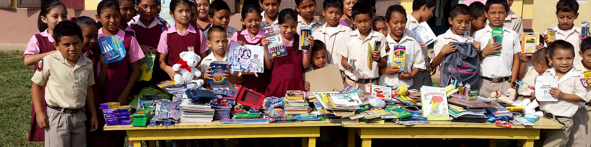 A group of children in school uniforms stands behind a table filled with books and school supplies outside a building.