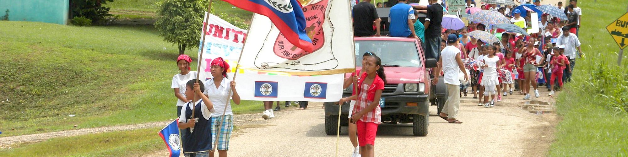 A group of people, including children, are marching down a rural road holding flags and banners, followed by cars and more participants.