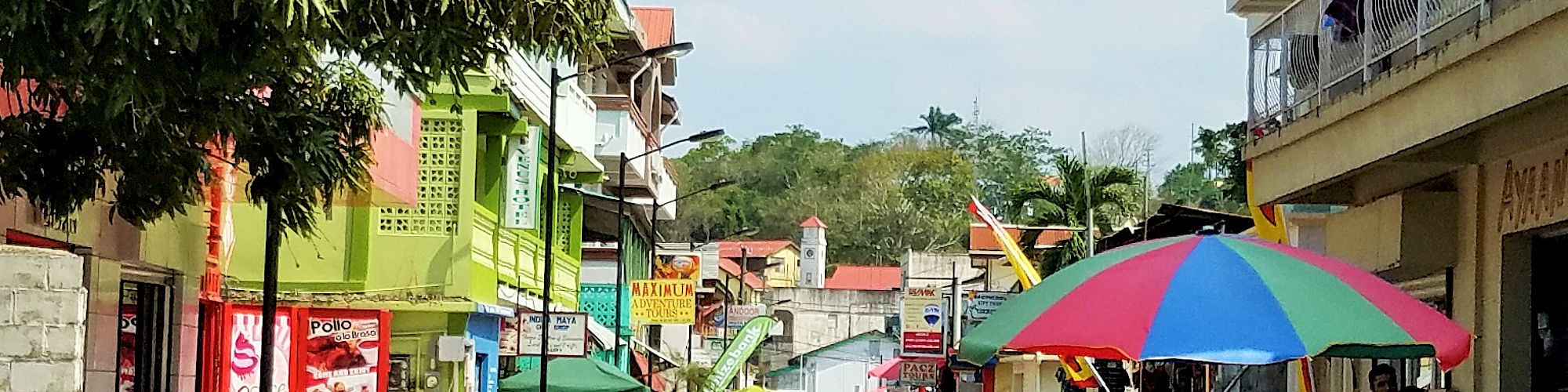 A lively street scene with colorful buildings, street vendors under umbrellas, and people walking and shopping, in a tropical setting.