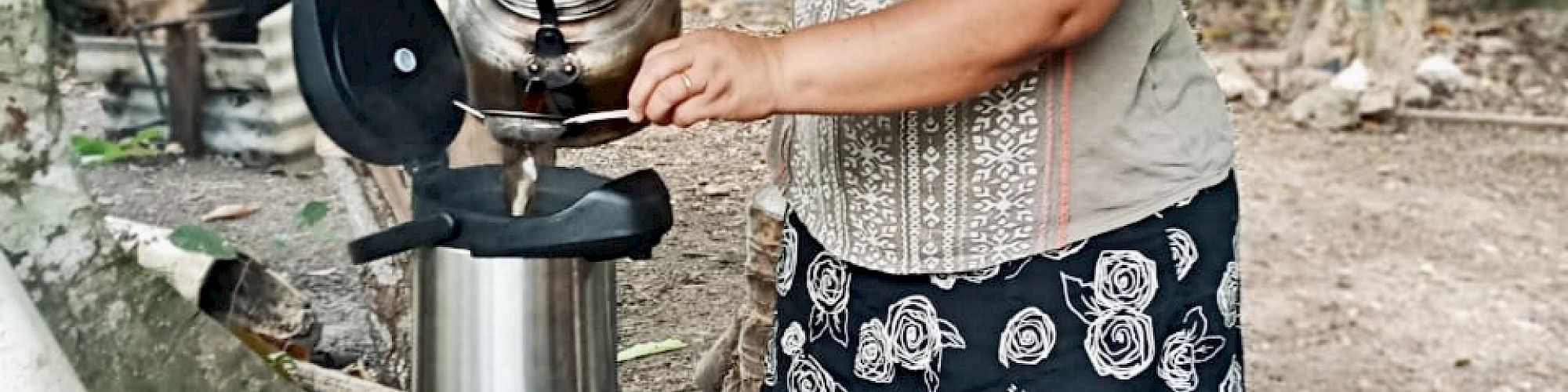 A person is outdoors using an outdoor kitchen setup, pouring water into a cooking pot beside other utensils, with trees and greenery in the background.