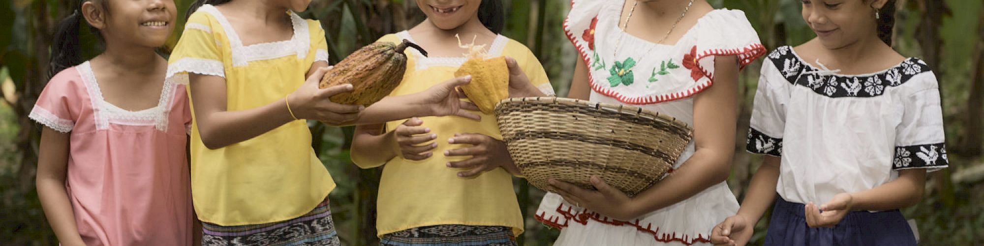 Five young girls in traditional clothing stand together outdoors, holding a cacao pod and a woven basket, smiling at each other.