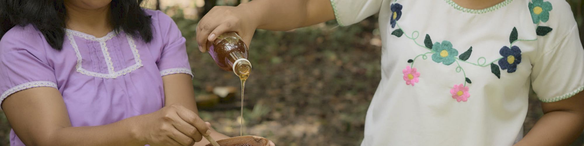 Two women are preparing food outdoors. One is holding a bowl, while the other pours a liquid into it. There is a stone grinder on the table.