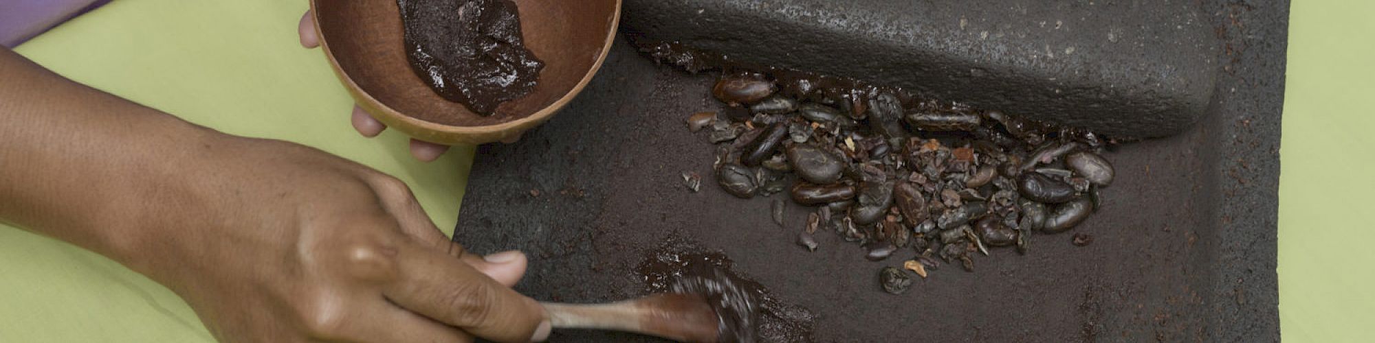 A person is grinding cocoa beans on a traditional stone slab, using a brown bowl and a grinding tool to make a chocolate paste.