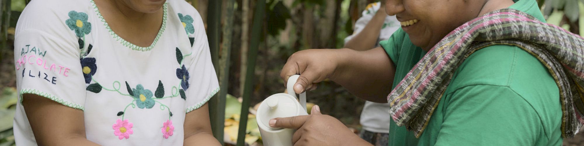 Two people are outdoors, with one pouring a liquid into a bowl held by the other; both are smiling and appear to be sharing a moment.