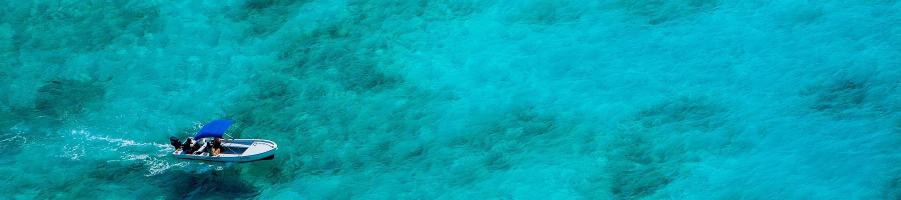 An aerial view of a small boat with people on board navigating clear, turquoise waters, with visible underwater features creating patterns in the sea.