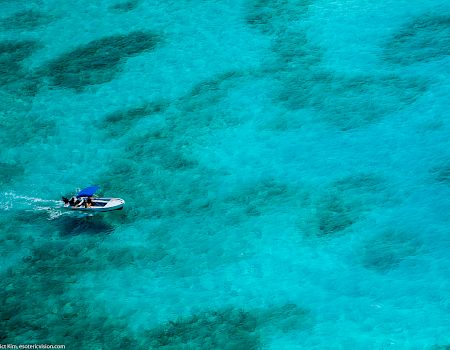 An aerial view of a small boat with people on board navigating clear, turquoise waters, with visible underwater features creating patterns in the sea.