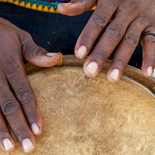 The image shows a close-up of hands playing a drum. The person wears a black garment with colorful patterns visible at the top.