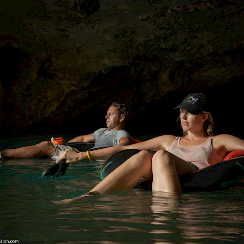 Two people are floating on tubes in a body of water inside a cave, one holding a beverage, both seeming relaxed.