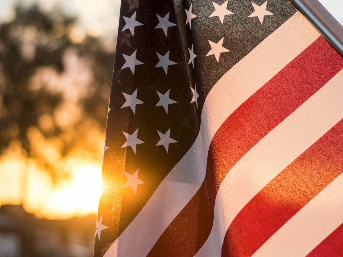 The image shows a close-up of the American flag with the sun setting in the background, casting a warm glow on the flag and the surrounding area.