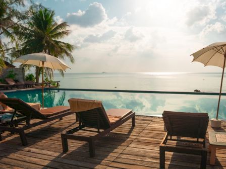 The image shows a tranquil poolside scene with lounge chairs, umbrellas, and a scenic view of the ocean, framed by palm trees and a clear sky.
