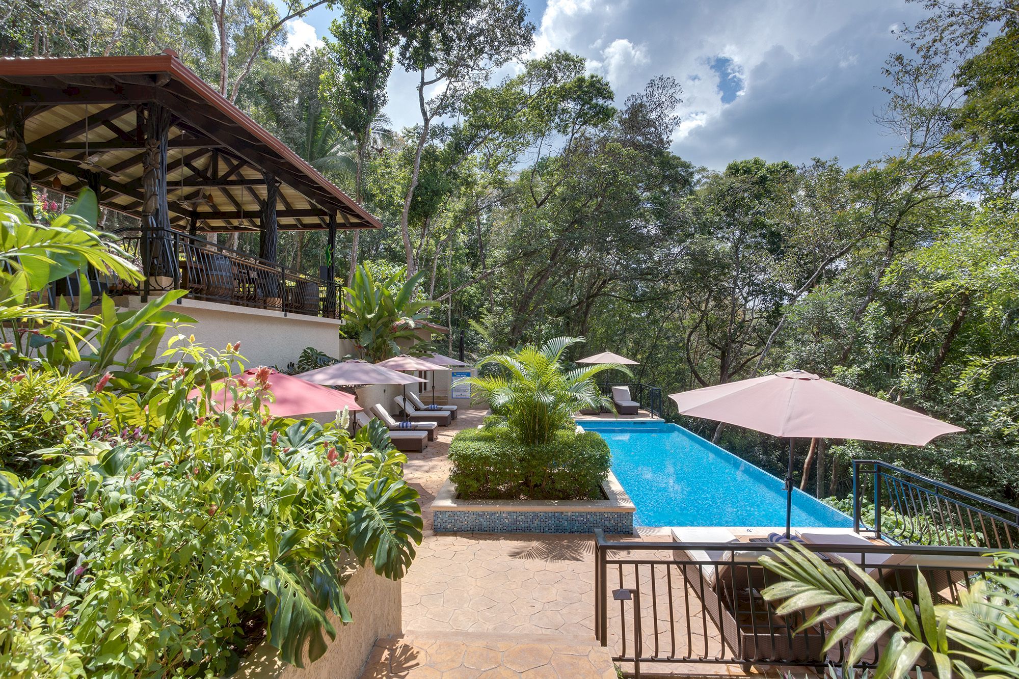 This image depicts a scenic outdoor area with a swimming pool, lounge chairs, umbrellas, lush greenery, and a covered seating area overlooking the pool.