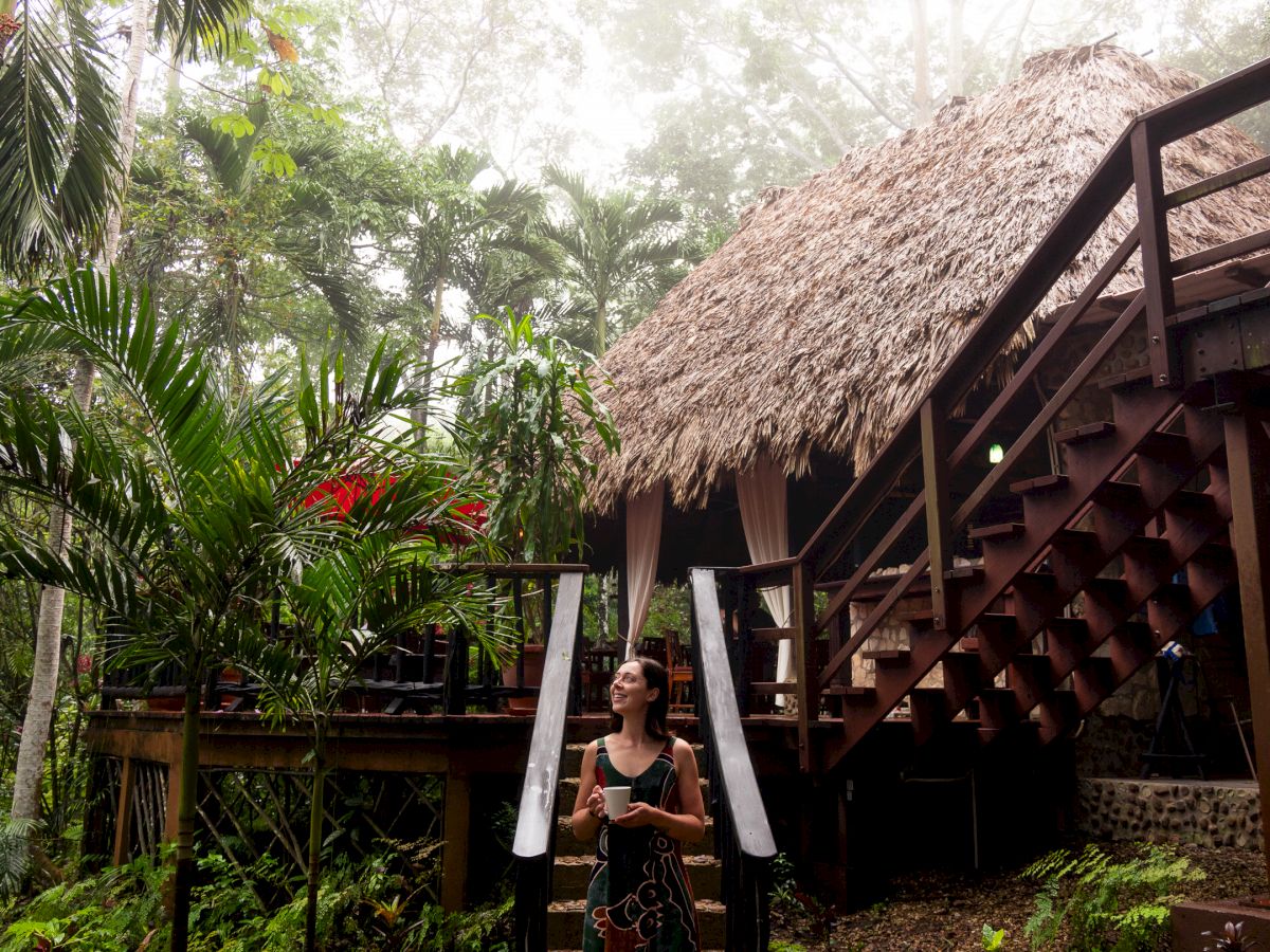 A person stands in front of a thatched-roof hut surrounded by lush greenery and palm trees, possibly in a tropical rainforest setting.