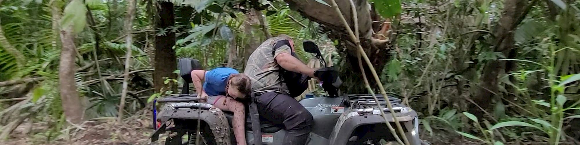 Two people on an ATV navigate through dense jungle terrain under a large fallen tree branch.