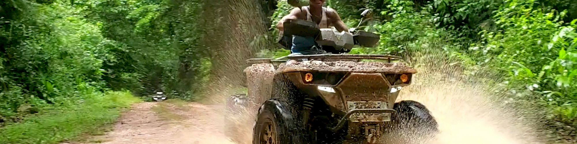 A person rides an ATV through a muddy puddle on a forest trail, causing a splash.