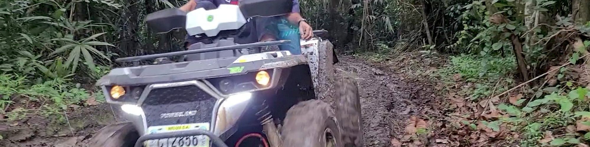 Two people are riding an ATV through a muddy forest trail.