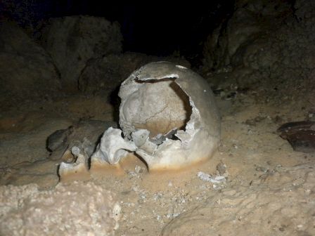 The image shows an old, broken skull lying on a sandy ground in what appears to be a cave.