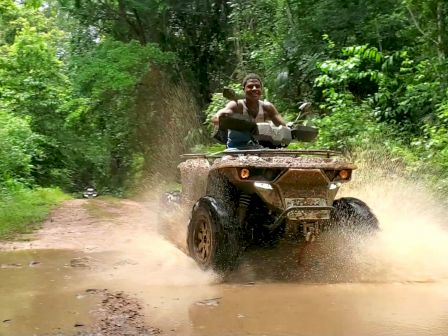 A person rides an ATV through a muddy puddle on a forest trail, creating a splash.