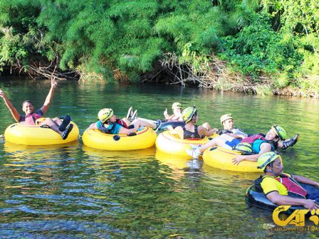 People in helmets and life vests are floating on yellow inner tubes in a river surrounded by green foliage, appearing to enjoy a tubing activity.