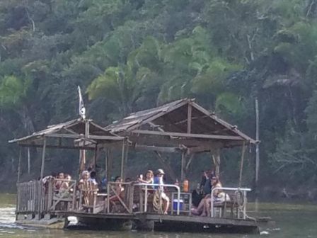 A group of people is gathered on a wooden raft in the middle of a body of water with a green forest in the background.