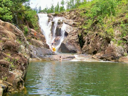 A picturesque waterfall with people swimming and jumping into a natural pool surrounded by rocky cliffs and greenery.