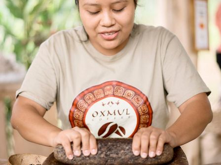 A person is using a traditional stone tool to grind something. The person is wearing a beige shirt with a round red logo.